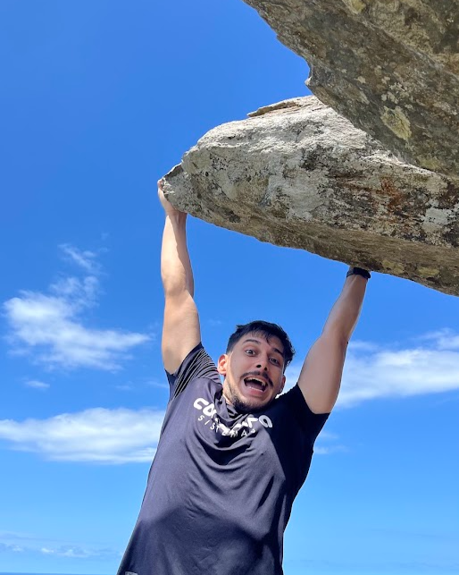 Ian Mateus Alves em perspectiva forçada, com os braços erguidos como se segurasse uma pedra enorme contra o céu azul